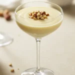 Close-up of a stemmed glass filled with a light yellow creamy drink, topped with chopped nuts, showcasing part of a Lotus Seed Honey Drink Recipe, with a blurred bowl of nuts in the background.