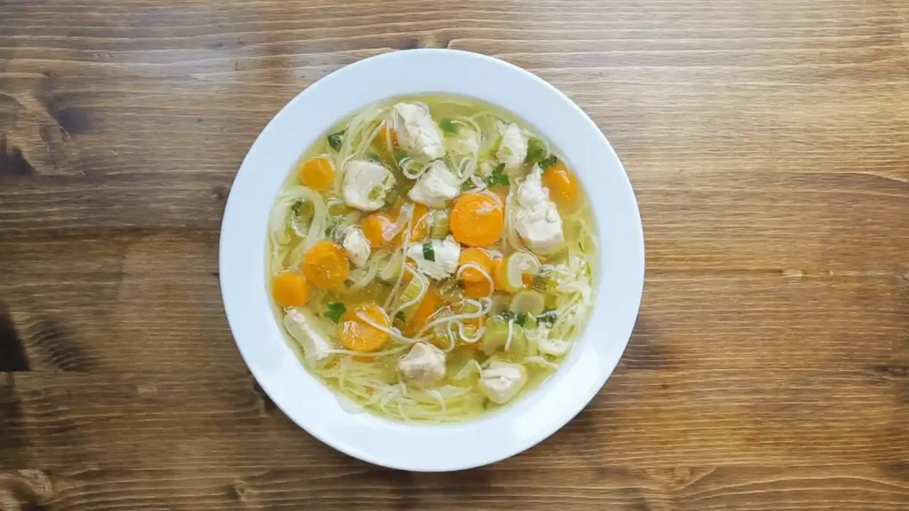 Overhead view of a comforting bowl of Chicken Bone Broth Noodle Soup with chicken, carrots, celery, and noodles on a wooden table.