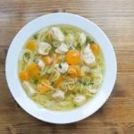 Overhead view of a comforting bowl of Chicken Bone Broth Noodle Soup with chicken, carrots, celery, and noodles on a wooden table.