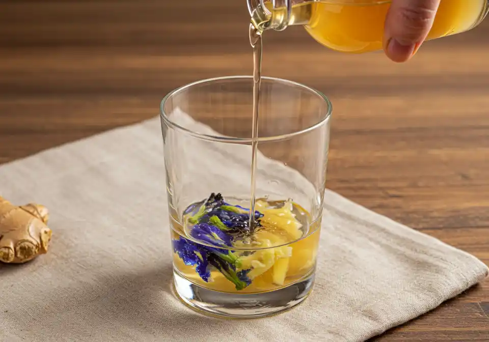 Pouring apple cider vinegar into blue butterfly pea flower infusion in a clear glass with grated ginger at the bottom on a linen cloth for Brazilian Mounjaro recipe preparation