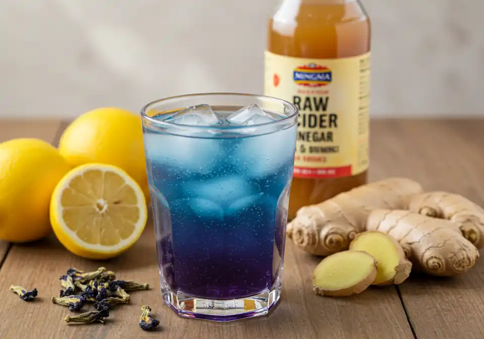 Clear glass of Brazilian Mounjaro recipe detox drink with vibrant blue-to-purple gradient served over ice and garnished with lemon wedge and ginger slice on a soft beige linen cloth